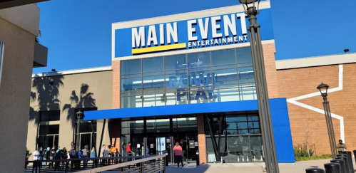 Exterior of Main Event Entertainment building with a clear blue sky and people waiting outside.