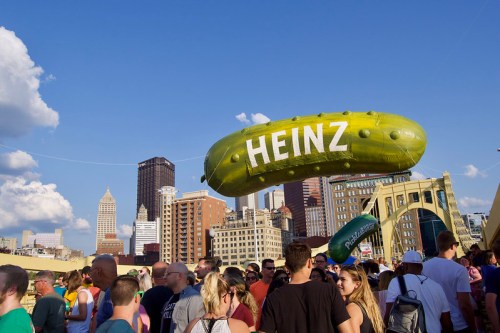 A large inflatable pickle with "HEINZ" floats above a crowd on a city bridge, with skyscrapers in the background.