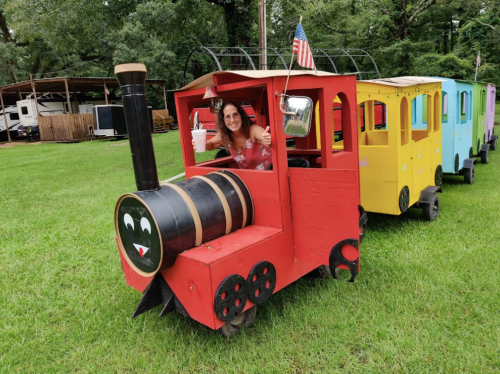 A woman gives a thumbs up from a colorful, wooden train in a grassy area, with trees in the background.