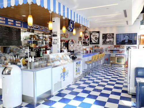 A retro-style diner with a checkered floor, counter seating, and vintage decor, featuring an ice cream display and a TV.