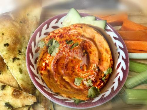 A bowl of hummus topped with olive oil and spices, surrounded by fresh vegetables and pita bread.