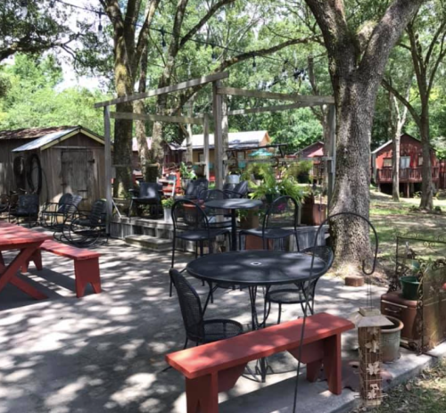 Outdoor seating area with tables and chairs surrounded by trees and rustic buildings.