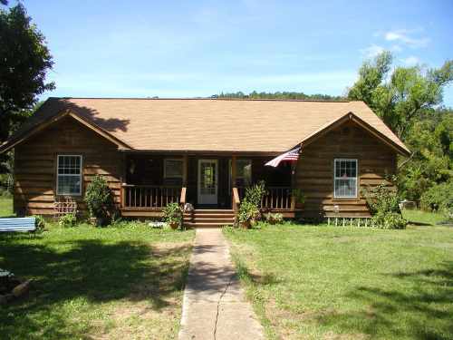 A rustic wooden house with a front porch, surrounded by greenery and a clear blue sky. An American flag is displayed.