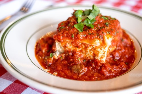 A slice of lasagna topped with marinara sauce and fresh basil, served on a white plate with a red checkered tablecloth.