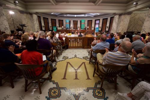 A crowded room with people seated, facing a panel at the front, featuring a decorative tiled floor with an "M" design.