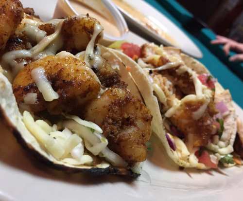 Close-up of two shrimp tacos topped with cabbage, tomatoes, and cheese, served on a plate with a side of sauce.