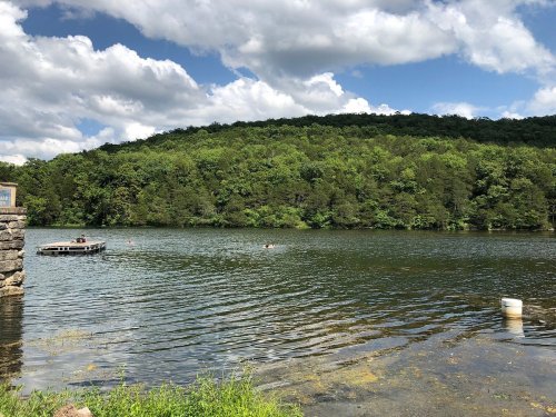 A serene lake surrounded by lush green hills under a partly cloudy sky, with a small dock visible in the foreground.