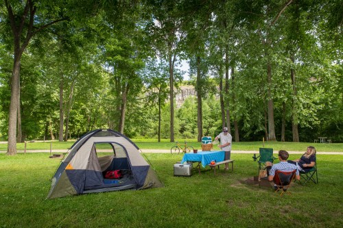 A campsite with a tent, picnic table, and people gathered around a fire in a lush green park.