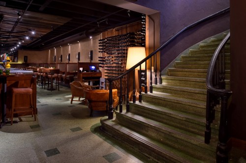 A dimly lit restaurant interior featuring a staircase, bar area, and wine display on the wall. Cozy seating is visible.