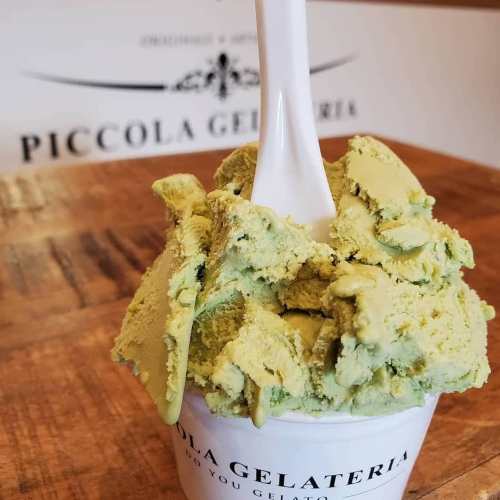 A scoop of green gelato in a cup with a white spoon, placed on a wooden surface.