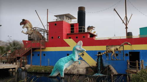 A colorful ship with dinosaur sculptures, including a T-rex and a blue dinosaur, set in a whimsical outdoor scene.