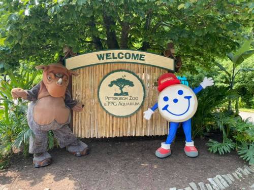 Two mascots stand beside a "Welcome" sign at the entrance of the Pittsburgh Zoo & PPG Aquarium, surrounded by greenery.