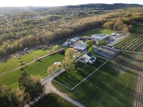 Aerial view of a rural farm with multiple buildings, green fields, and trees in the background under a clear sky.