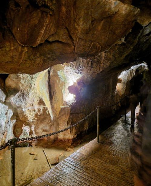 A dimly lit cave interior with rocky walls and a chain barrier along a wet path. Light filters in from an opening.