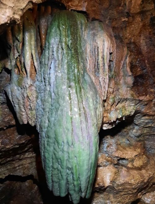 A green, elongated stalactite hanging from a cave ceiling, surrounded by rocky formations.