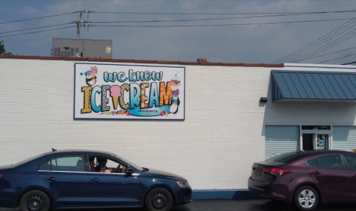 A colorful sign on a building reads "We Know Ice Cream" with a blue sky and parked cars in front.