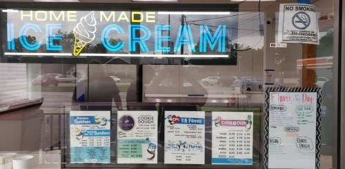 A storefront window displaying a neon sign for homemade ice cream and various menu items and prices.