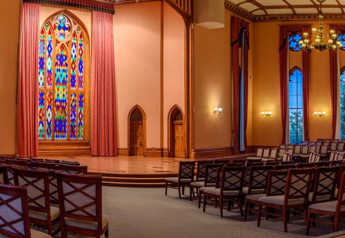 Interior of a hall featuring stained glass windows, wooden stage, and rows of chairs arranged for an event.