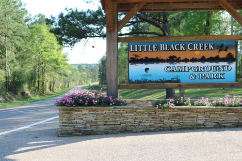 Sign for Little Black Creek Campground & Park, surrounded by flowers and trees, with a road leading into the distance.