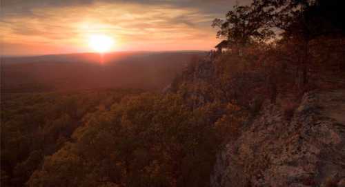 A sunset over a vast landscape, with trees and a rocky cliff in the foreground.