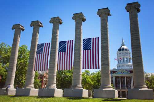 Five tall columns with an American flag draped between them, set against a blue sky and a historic building in the background.