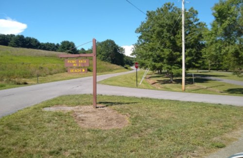 A wooden sign at a road intersection indicating directions to a picnic area and beach, surrounded by greenery.