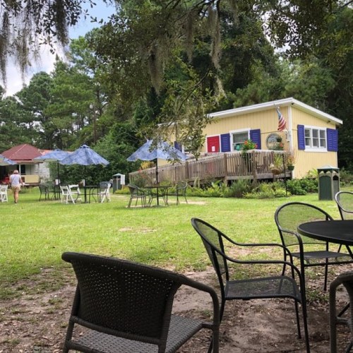 A grassy area with chairs and tables, featuring a yellow building with blue shutters and an American flag in the background.
