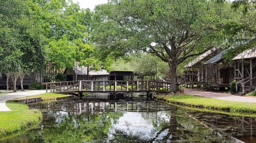 A wooden bridge crosses a calm pond surrounded by lush greenery and rustic buildings in a serene outdoor setting.