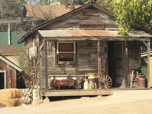 A rustic wooden house with a metal roof, porch, and vintage farm equipment in a rural setting.