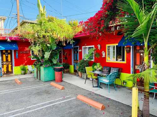 Colorful building with red walls, vibrant plants, and outdoor seating in a sunny parking area.