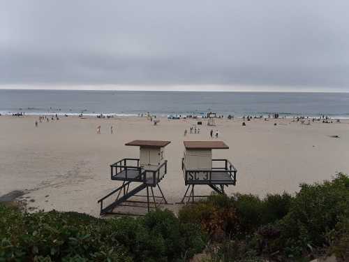 Two lifeguard towers on a sandy beach with people enjoying the shore under a cloudy sky.