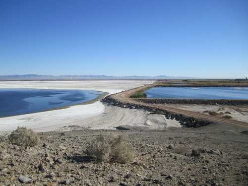 A landscape featuring salt flats and blue water bodies under a clear blue sky, with distant mountains in the background.