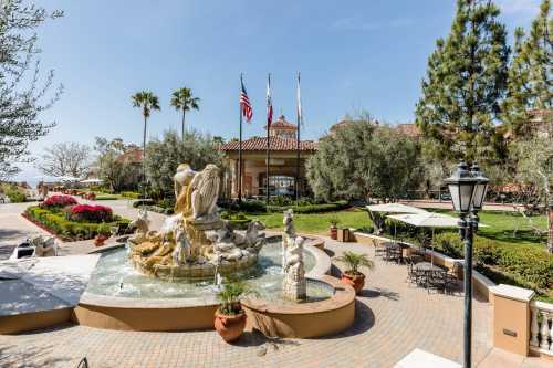 A scenic view of a fountain surrounded by greenery and palm trees, with a building and flags in the background.