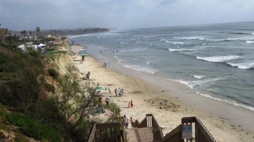 A sandy beach with gentle waves, people walking, and cliffs in the background under a cloudy sky.