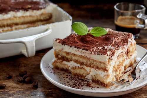 A slice of tiramisu on a plate, garnished with mint, next to a glass of coffee and a baking dish in the background.