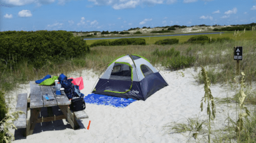 A tent set up on a sandy beach, with a picnic table nearby and lush greenery in the background under a blue sky.