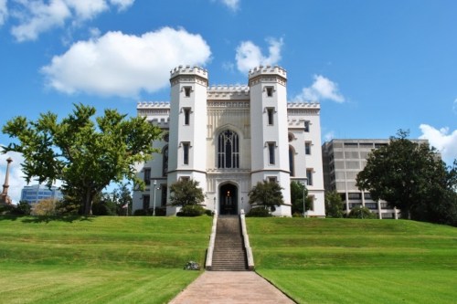 Historic castle-like building with towers, surrounded by green grass and trees, under a blue sky with clouds.