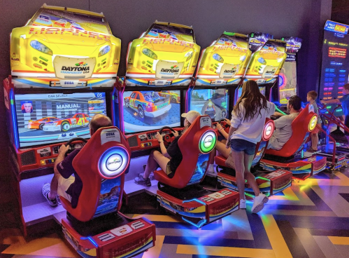 A group of people playing racing arcade games in a colorful gaming area.