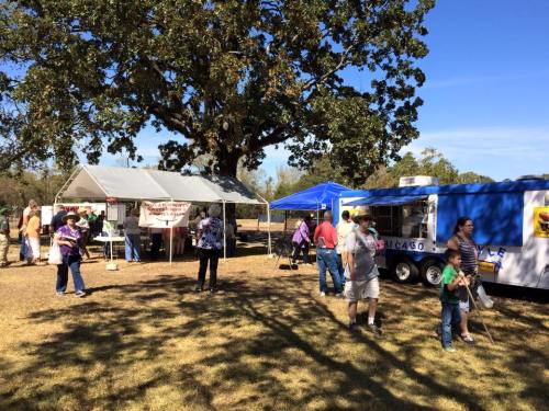 A sunny outdoor scene with food stalls, people walking, and a large tree in a park setting.