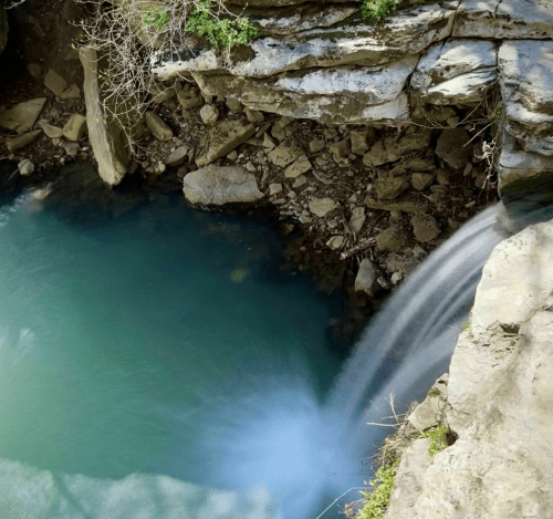 A serene waterfall cascading into a clear blue pool surrounded by rocky cliffs and sparse vegetation.