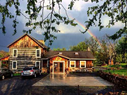 A rustic wooden house with a rainbow in the background, surrounded by greenery and a cloudy sky.