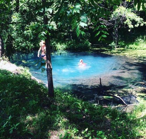 A serene blue pond surrounded by greenery, with one person swimming and another standing nearby.