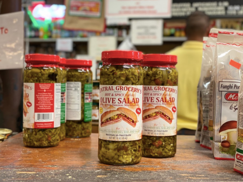 Jars of Central Grocery's hot and spicy olive salad on a wooden counter, with snacks in the background.