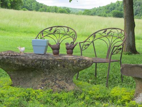 A rustic stone table with two decorative metal chairs and potted plants, set in a grassy field with trees in the background.