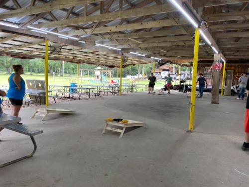 A spacious outdoor pavilion with picnic tables and people playing cornhole games. Bright lights illuminate the area.