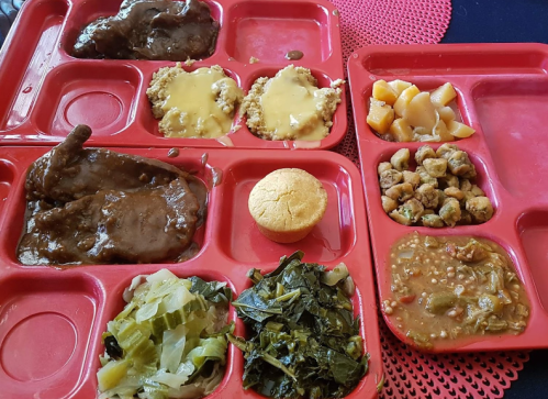 A red cafeteria tray with two servings of meat, cornbread, vegetables, and fruit in separate compartments.