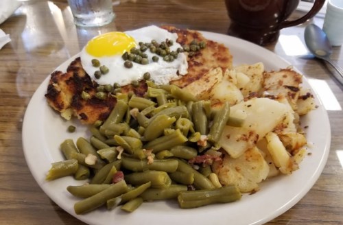A plate of food featuring a fried egg, capers, hash browns, green beans, and diced potatoes.