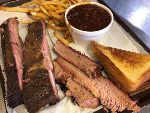 A plate featuring smoked brisket, ribs, baked beans, French fries, and toasted bread on a tray.