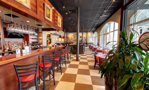 Interior of a restaurant featuring a bar, tables, and large windows, with a warm, inviting atmosphere.