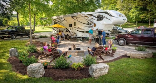 A group of people enjoying a gathering around a fire pit near an RV, with trees and greenery in the background.